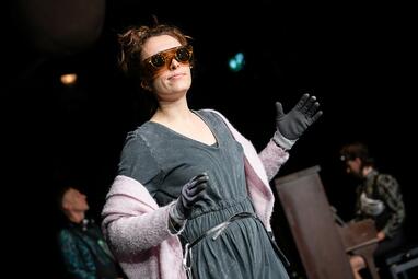 Vergrößerte Ansicht von Dunkle Bühne: Eine Schauspielerin in grauem Overall und mit rosaner Wolldecke über den Schultern trägt zwei Brillen übereinander