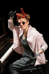 Vergrößerte Ansicht von Eine Schaupielerin sitzt am Klavier, mit geekeltem Gesichtausdruck fühlt sie nach der braunen Kackwurst, die gerade auf ihrem Kopf landet.