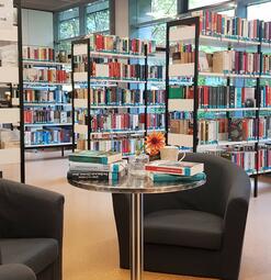 Vergrößerte Ansicht von Foto der Zweigstelle Herzogenried. Man sieht zwei dunkle Sessel, davor einen kleinen runden Tisch mit Büchern und einer Blume. Im Hintergrund sieht man Bücherregale.