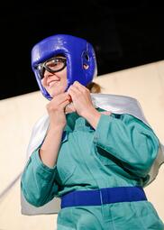 Vergrößerte Ansicht von Marie-Tschann Kromminga trägt einen Kopfschutz fürs Boxen sowie eine Schutzbrille. Sie bindet sich ein kleines Cape um.
