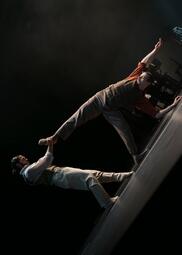 Vergrößerte Ansicht von Ein Tänzer hält einen anderen Tänzer am Fuss fest, während dieser akrobatisch nach hinten fällt.