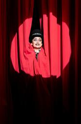 Vergrößerte Ansicht von Annemarie Brüntjen reckt grinsend mit aufgerissenen Augen ihren Kopf zwischen den roten Bühnenvorhängen hervor.
