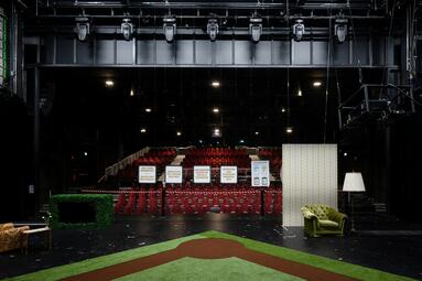 Vergrößerte Ansicht von Blick von der Bühne in den Publikumsraum. Aus dem Bühnenboden liegt ein Teppich im Stil eines baseballfeldes sowie verschiedene sessel drum herum. Der Bühnenrand ist mit einem Absperrseil versehen. Zwischen den Sitzreihen stehen schilder mit…