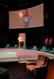 Vergrößerte Ansicht von Das Bühnenbild mit baseball-Rasen und einem Sessel davor. Um den Teppich befinden sich Kugeln im gleichmäßigen Abstand, überdeckt mit einem grünen Tuch. Auf einer Leinwand über der Bühne erscheint eine projektion eines Gartenzwerges, welcher in…
