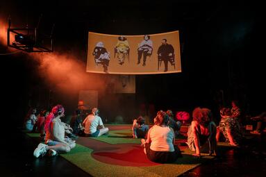 Vergrößerte Ansicht von Das Publikum trägt ein Headset und schaut auf die Leinwand. Dort sind Ausschnitt von vier sitzenden Personen auf orangenen Hintergrund abgebildet.