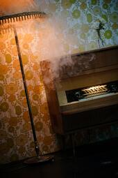 Vergrößerte Ansicht von Eine rötlich leuchtende Lampe und ein Klavier. Von der Lampe dringt Nebel.