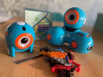 Vergrößerte Ansicht von Ein Foto der kugelförmigen Mini-Roboter Dash und Dot mit Legobausteinen. Ihr Gehäuse ist hellblau, der Ring um ihre schwarze Kameralinse leuchtend orange.