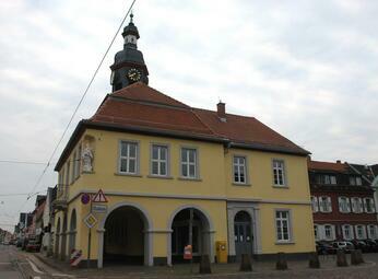 Vergrößerte Ansicht von Ein Foto zeigt das Rathaus von Seckenheim unter bewölktem Himmel. Das zweigeschossige Gebäude hat eine gelbe Fassade mit hellgrau gerahmten Sprossenfenstern und Rundbögen im Erdgeschoss.