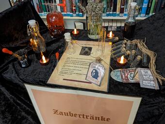 Vergrößerte Ansicht von Ein Foto von einem mit schwarzem Samt umhüllten Tisch vor einem Bücherregal. An der Stirnseite hängt ein Plakat mit dunkelroter Schrift auf blassrosa Hintergrund: „Zaubertränke“. Auf dem Tisch liegt ein Rezept für den „Trank der lebenden Toten“. Um das Rezept herum Flaschen mit dunkler Flüssigkeit, Pipetten, Erlenmeyerkolben, leere Reagenzgläser und Teelichter.