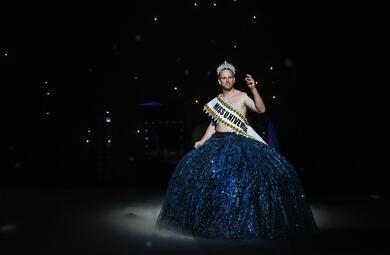 Vergrößerte Ansicht von Eddie Irle trägt ein Diadem und einen breiten, blau-glitzernden Rock. Über seinen Oberkörper trägt er einen Banner mit der Aufschrift &quot;Miss Universe&quot;. Er hebt eine Hand.