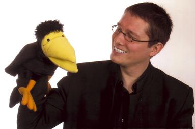 Vergrößerte Ansicht von Auf einem Foto neigt TINO seinen Kopf zu einer Raben-Handpuppe aus Plüsch. Tino dunkles Haar ist kurz. Er trägt eine Brille mit schmalen ovalen Gläsern und ein schwarzes Hemd. Die Raben-Handpuppe hat schwarze Knopfaugen, einen großen gelben Schnabel und orangefarbene Beine und Krallen.