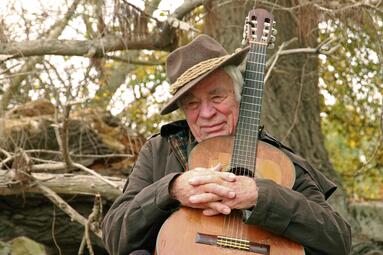 Vergrößerte Ansicht von Auf einem Foto sitzt der weißhaarige Alfred Baumgartner vor einem Baum mit dickem Stamm und hält eine Gitarre umschlungen. 