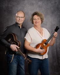 Vergrößerte Ansicht von Auf einem Portraitfoto stehen ein Mann mit Halbglatze und Brille und eine Frau mit kinnlanger Dauerwelle nebeneinander. Lächelnd halten sie Ukulelen hoch.