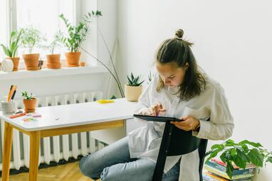 Vergrößerte Ansicht von Ein jugendliches Mädchen sitzt in einem hellen Raum mit Pflanzen und hat ein Tablet in der Hand, auf das sie schaut.