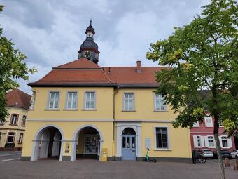 Vergrößerte Ansicht von Ein Foto zeigt das Rathaus von Seckenheim unter bewölktem Himmel. Das zweigeschossige Gebäude hat eine gelbe Fassade mit hellgrau gerahmten Sprossenfenstern 