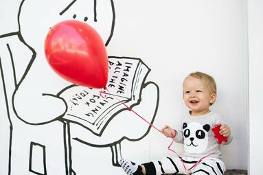 Vergrößerte Ansicht von Ein Kleinkind sitzt lachend vor einer Wand mit Comic-Motiv und hält einen roten Luftballon an der Schnur.