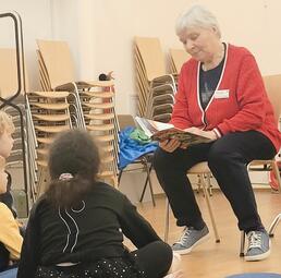 Vergrößerte Ansicht von Eine Frau sitzt auf einem Stuhl und liest aus einem Buch, vor ihr sitzen Kinder auf dem Boden.