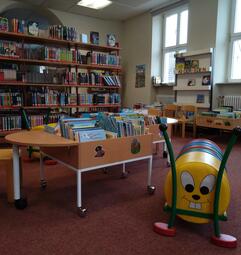 Vergrößerte Ansicht von Ein Foto aus dem Kinderbereich der Stadtteilbibliothek Seckenheim. Zwischen einem Regal mit Büchern und einem Sitzkissen-Halter in Raupenform stehen leicht versetzt zwei viereckige Tröge mit Bilderbüchern.