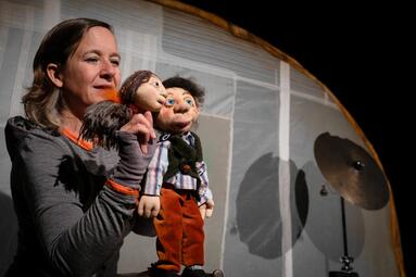 Vergrößerte Ansicht von Ein Foto zeigt Theatermacherin Maren Kaun mit zwei Figurenpuppen auf einer Bühne.