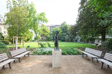 Vergrößerte Ansicht von Lameygarten: Platz mit vier Bänken, Reiter-Statue und frisch gepflanztem Baum. Im Hintergrund eine Wiese und ältere Bäume.