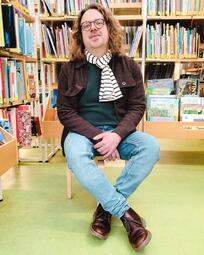 Vergrößerte Ansicht von Feuergriffelpreisträger Markus Altmeyer - Jeans, braune Jacke, Brille, lange Haare - sitzt vor Bücherregalen in der Kinder- und Jugendbibliothek