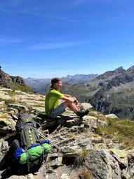 Vergrößerte Ansicht von Auf einem Foto sitzt Frank Eichmann auf dem felsigen Boden eines Gebirgskamms im Sonnenschein. Frank blickt in die Ferne und lächelt. Er trägt ein Käppi, kurze Funktionskleidung und Wanderstiefel. Neben ihm liegt sein großer Wanderrucksack auf dem Boden.