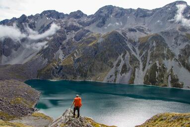 Vergrößerte Ansicht von Ein Blick aus der Ferne auf Jan Rumig. Er steht mit einem leuchtend orangefarbenen Wanderrucksack auf einem Felsvorsprung und schaut auf die glatte Oberfläche eines blaugrauen Bergsees hinab. Hinter dem See ragt eine schroffe Gebirgswand steil empor.