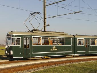 Vergrößerte Ansicht von Eine alte Straßenbahn - bedruckt mit dem Eichbaum-Bier-Symbol - fährt an einem sonnigen Tag auf Schienen.