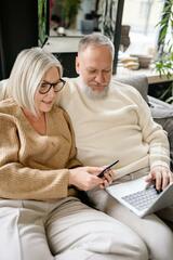 In einem Wohnzimmer sitzen ein Mann und eine Frau nebeneinander auf einem Sofa. Er blickt auf einen Laptop auf seinem Schoß, sie in ein Smartphone in ihrer Hand. Beide haben silbergraues Haar.